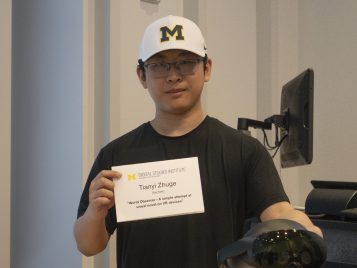 Tianyi Zhuge stands wearing a white University of Michigan hat , holding a black virtual reality headset and a paper displaying his name and project title.